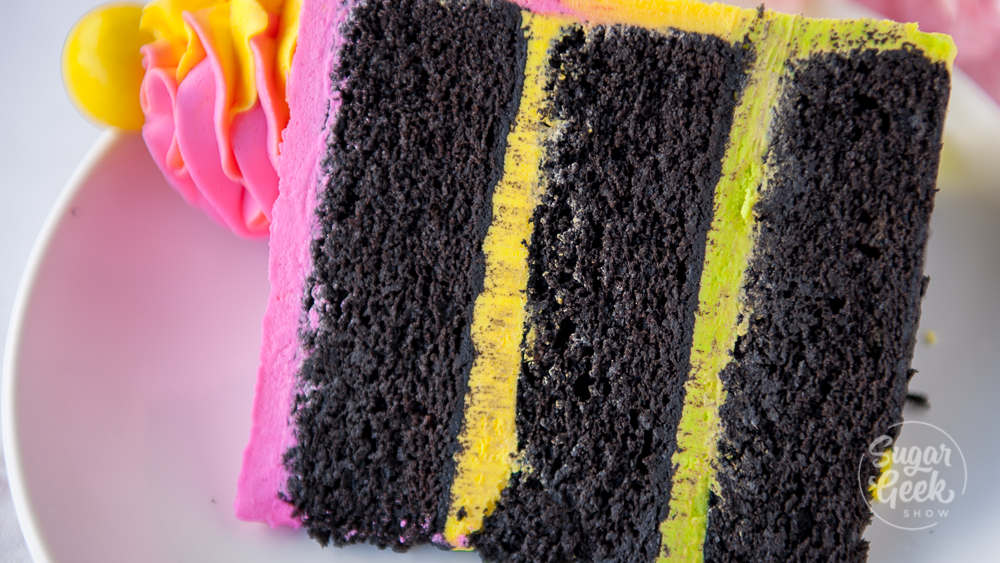 slice of black velvet cake with bright pink and yellow buttercream frosting on a white plate