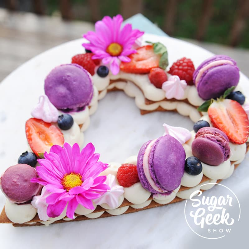 cream tart in the shape of a heart topped with fresh flowers, fruit and macarons