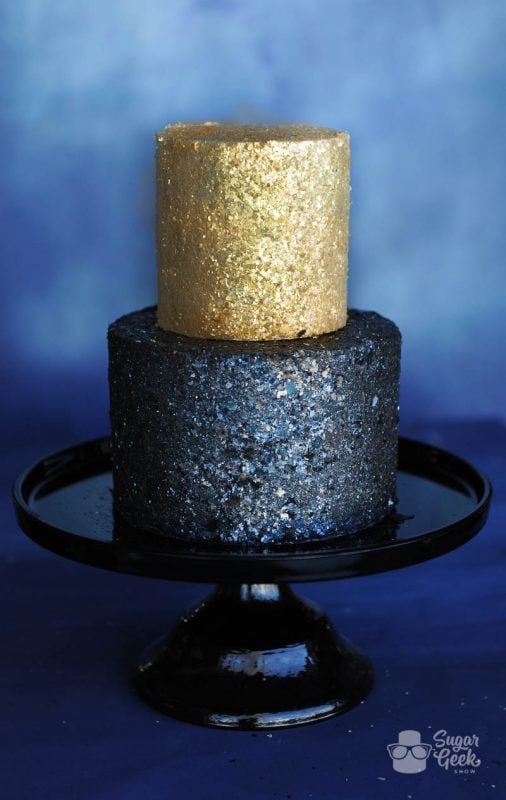 two tier cake covered in gold and black glitter on a black cake stand