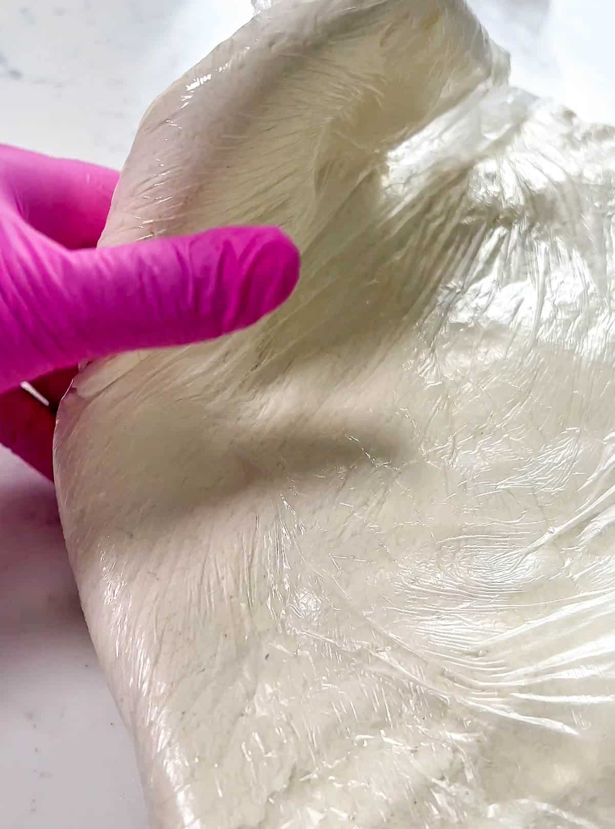 Pink gloved hand bending modeling chocolate wrapped in plastic wrap.