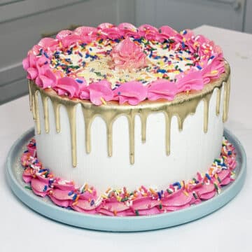 close up of a white cake with gold drip and pink piped borders on a blue plate