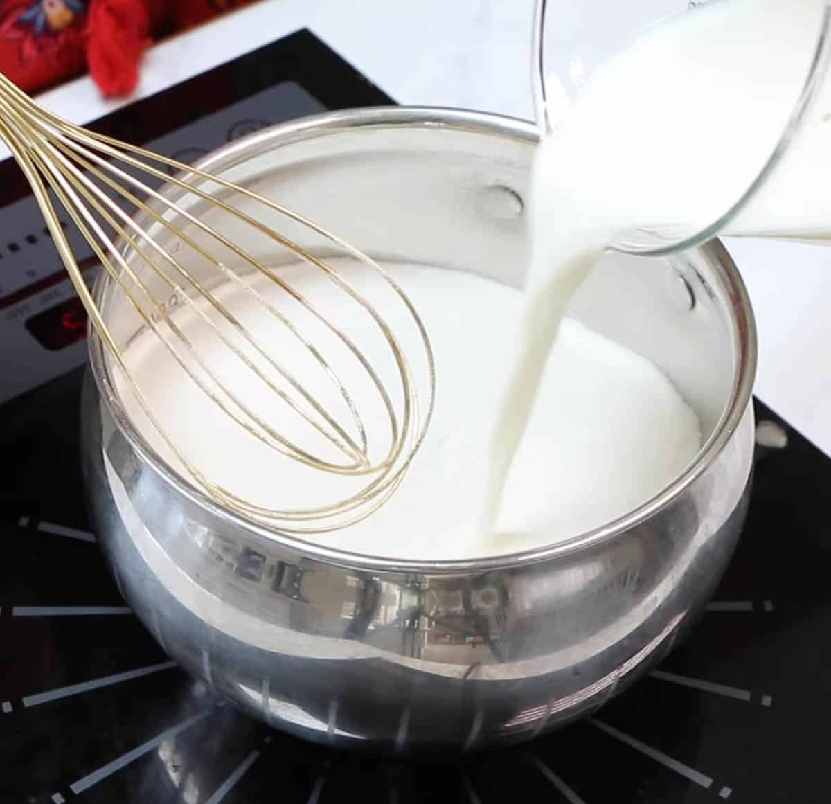 Pouring milk into a metal saucepan with flour and sugar and a whisk.