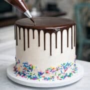chocolate drip being piped onto a white cake with rainbow sprinkles