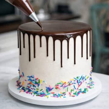 chocolate drip being piped onto a white cake with rainbow sprinkles