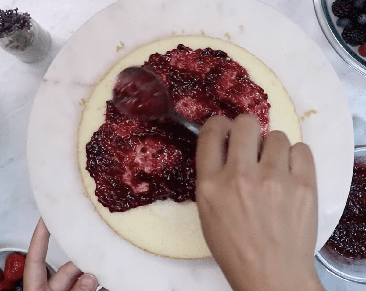 Hand spreading berry mixture on top of a vanilla cake layer
