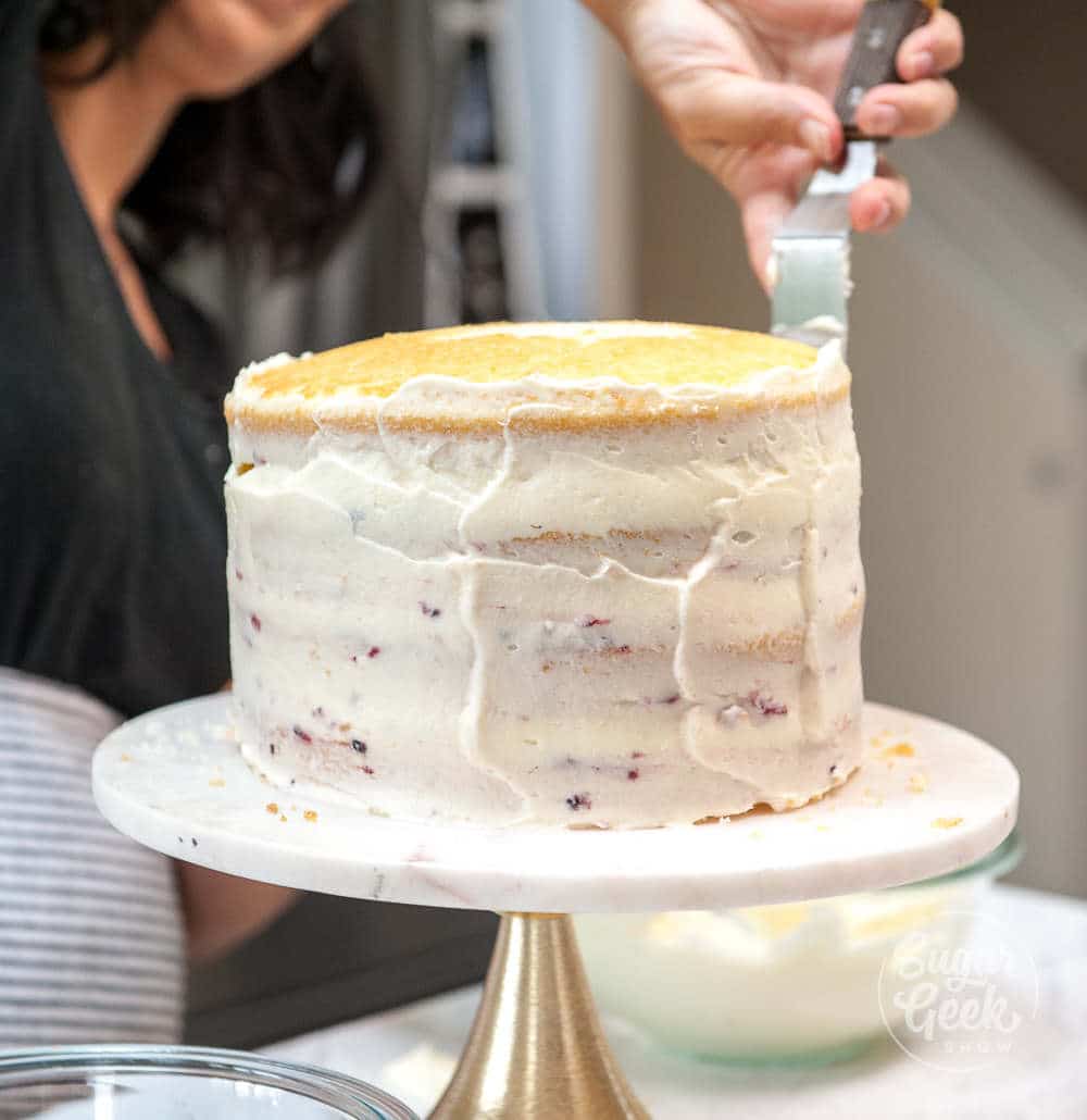 Cake on a cake pedestal being frosted with an offset spatula.