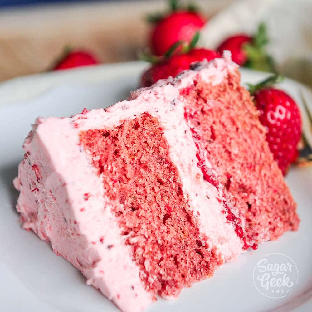 slice of fresh strawberry cake on a white plate with strawberries behind it