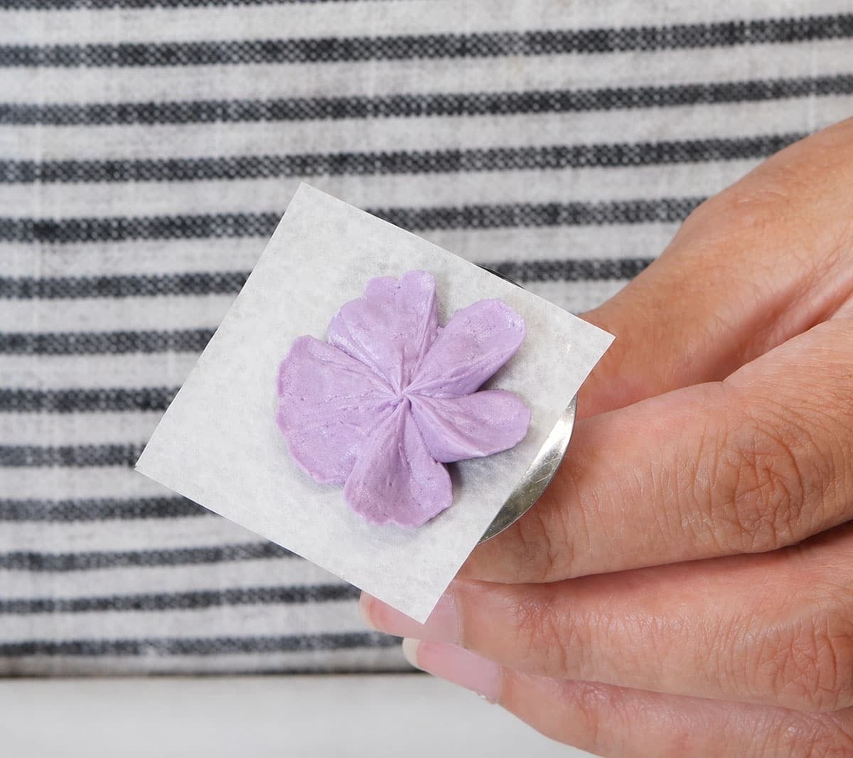 Buttercream flower on a parchment square on a buttercream nail held by a hand.