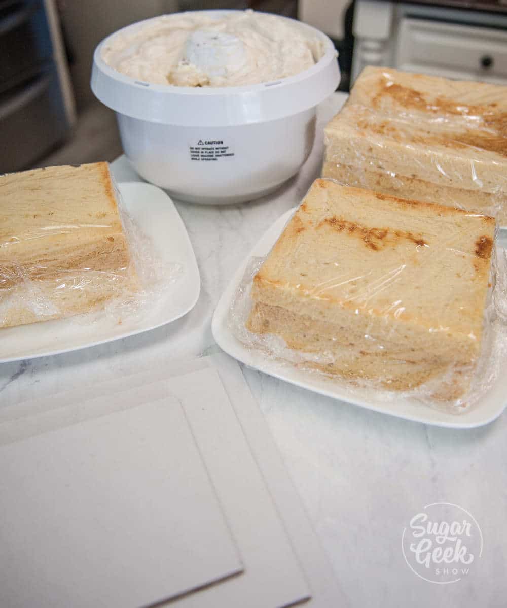 square cakes wrapped in plastic wrap with bowl of buttercream in background