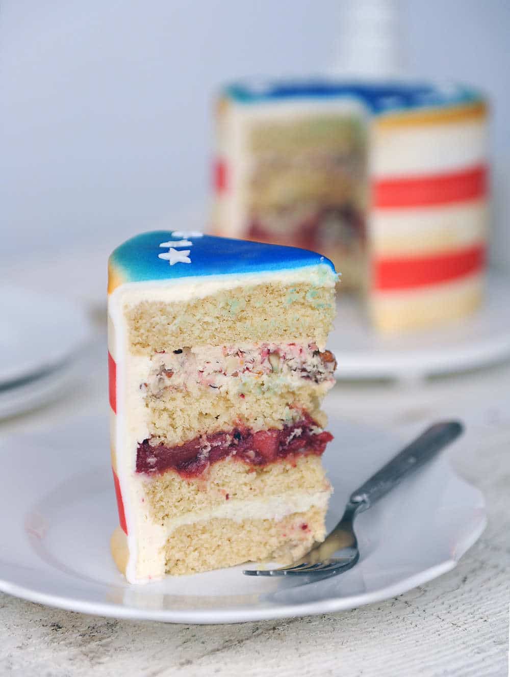 slice of brown butter cake with apple cranberry filling and vanilla bean buttercream on a white place. Fondant airbrushed on top blue with white stars. Red and white stripes around the sides