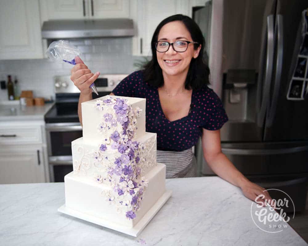 liz marek smiling at the camera in her kitchen holding a piping bag standing behind three tier square white wedding cake with purple buttercream flowers flowing down the front