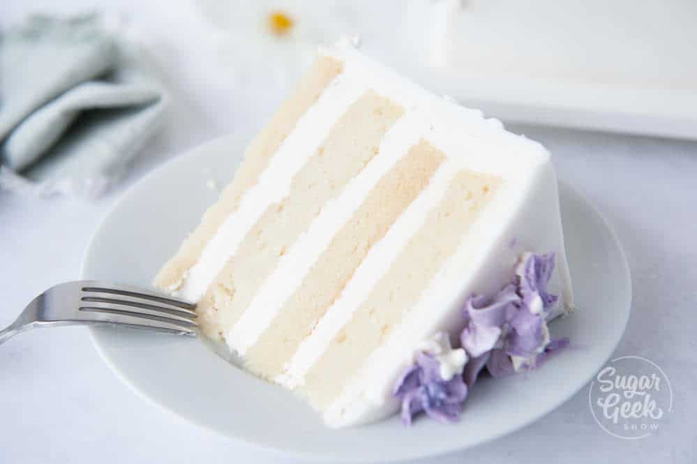 Slice of vanilla cake with buttercream and purple buttercream flowers on a white plate