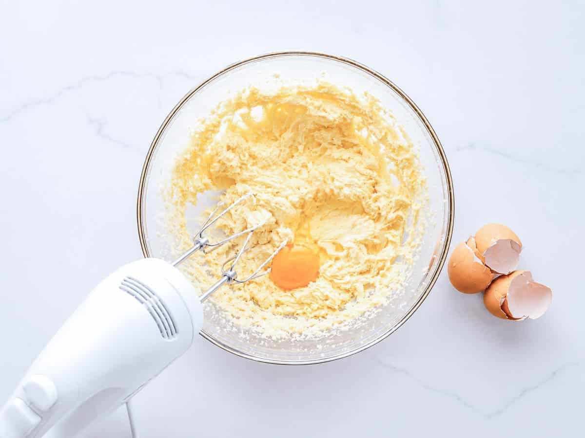 Hand mixer creaming eggs, butter, and sugar together in a glass bowl.