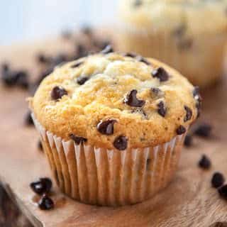 close up of moist chocolate chip muffin on wooden cutting board with blurry chocolate chip muffin in the background and loose chocolate chips all around the muffin