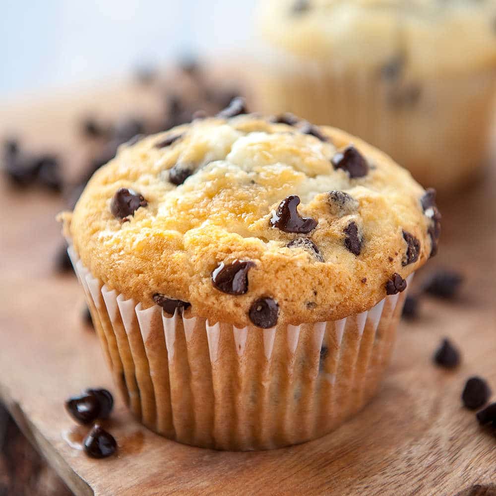 close up of moist chocolate chip muffin on wooden cutting board with blurry chocolate chip muffin in the background and loose chocolate chips all around the muffin