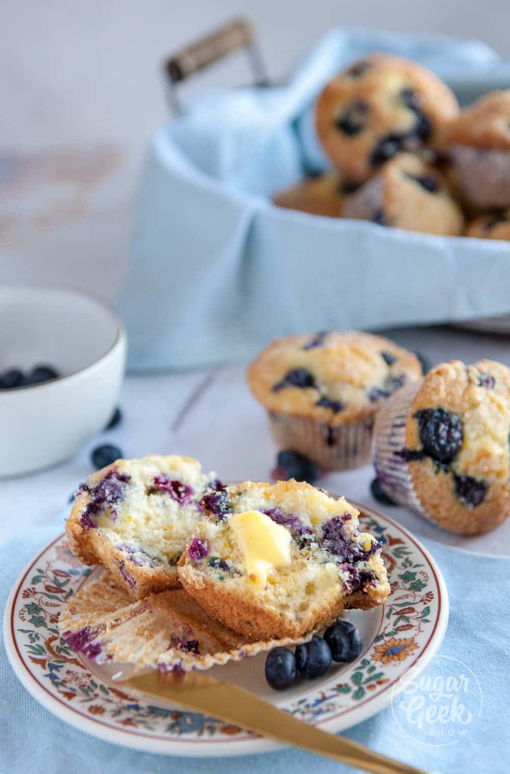 lemon blueberry muffin sliced open with melted butter. Sitting on plate with floral border and butter knife on the edge. Basket of muffins in the background.