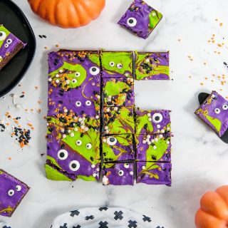 monster mash cheesecake brownies cut into squares on a white background. Sprinkles, pumpkins and plates around the brownies