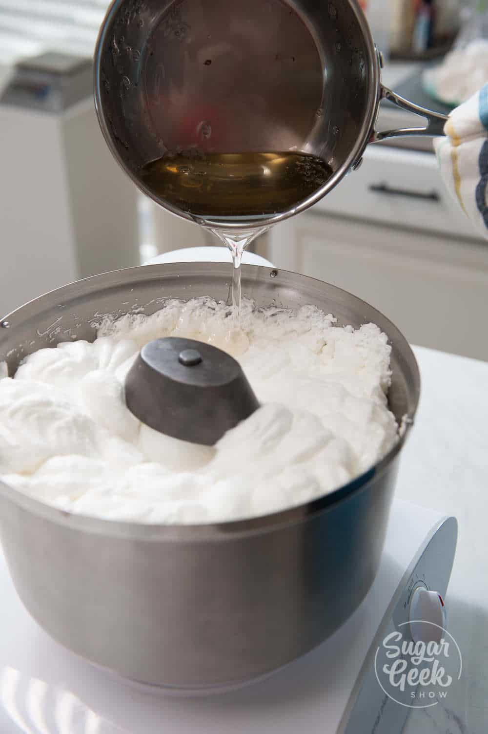 Metal saucepan pouring hot sugar into a mixer whipping egg whites.