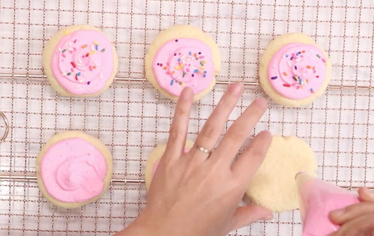 Piping pink American buttercream onto Lofthouse cookies on a cooling rack.