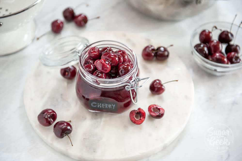 cherry filling in a glass jar on a white table with cherries surrounding it