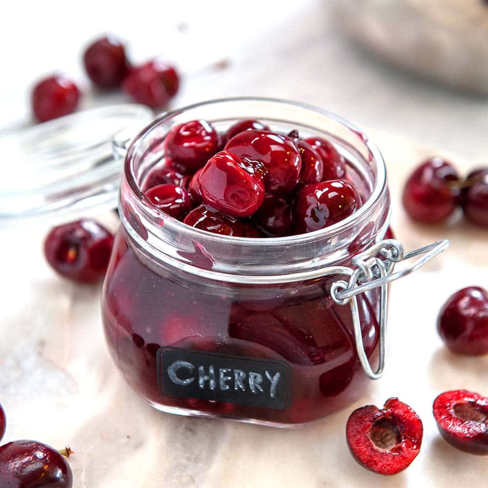 homemade cherry filling in a glass jar with the lid open. Black label with cherry written on the front. Cherries surrounding the jar on a white table
