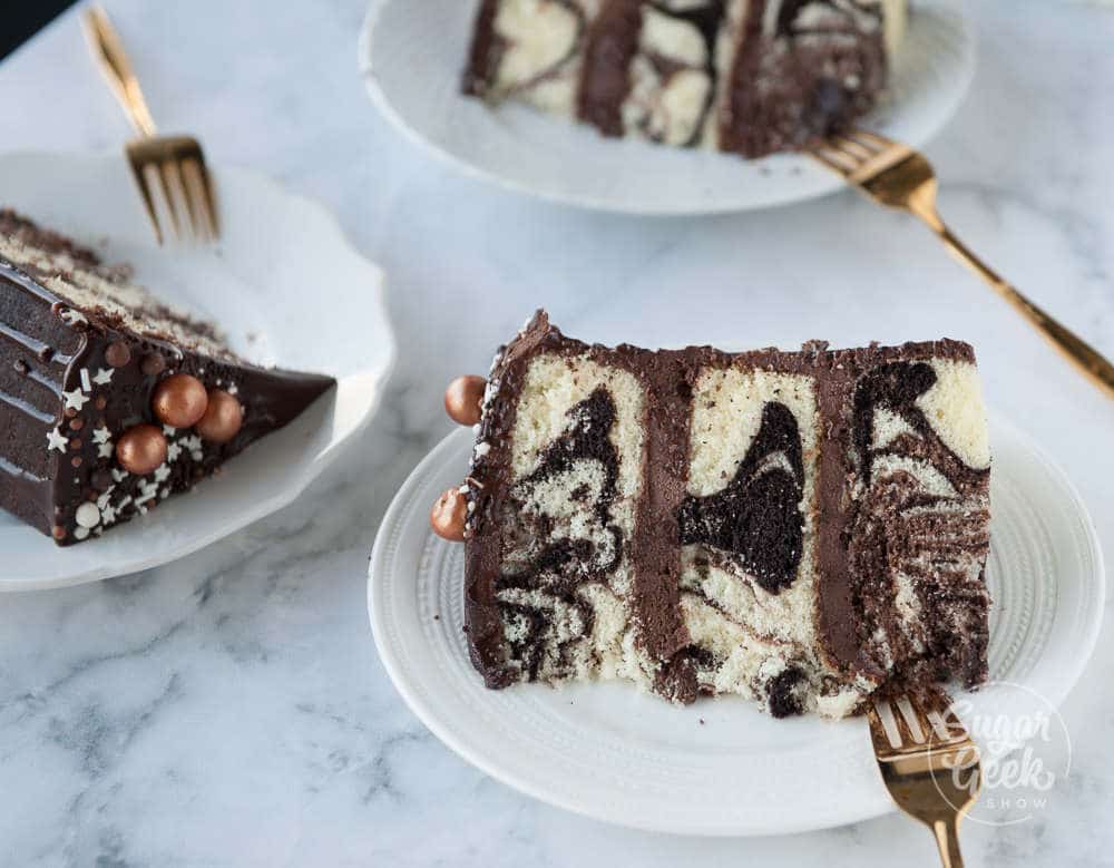 three slices of marble cake with chocolate frosting on white plates with gold forks