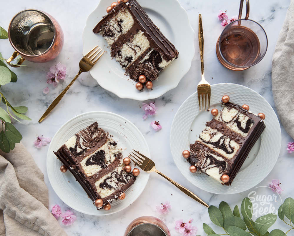 three slices of marble cake on three white plates with gold forks. Shot from above. Surrounded by three copper mugs, greenery and cherry blossoms