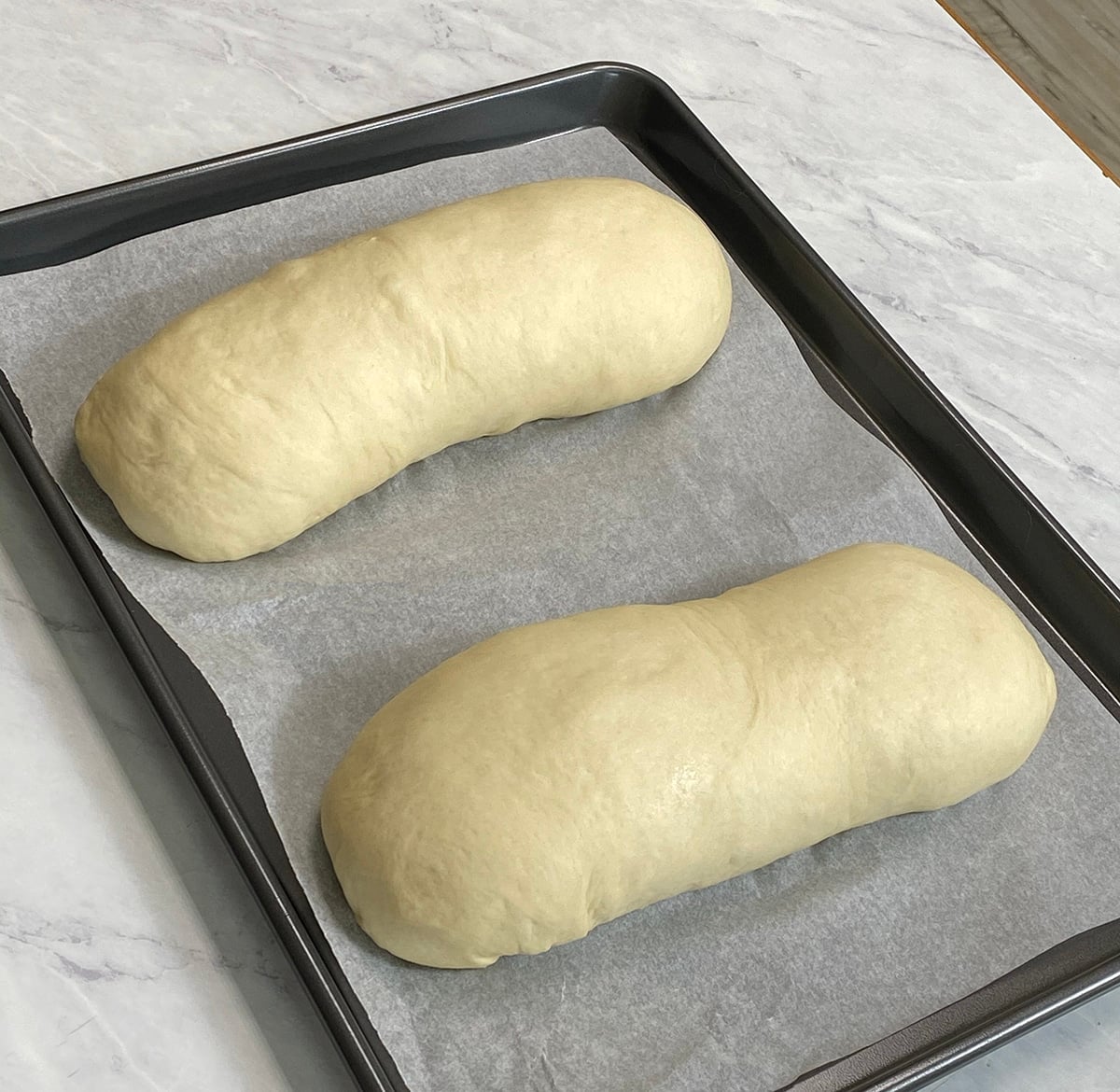 Two loaves of unbaked sandwich bread dough on a sheet pan lined with parchment paper.