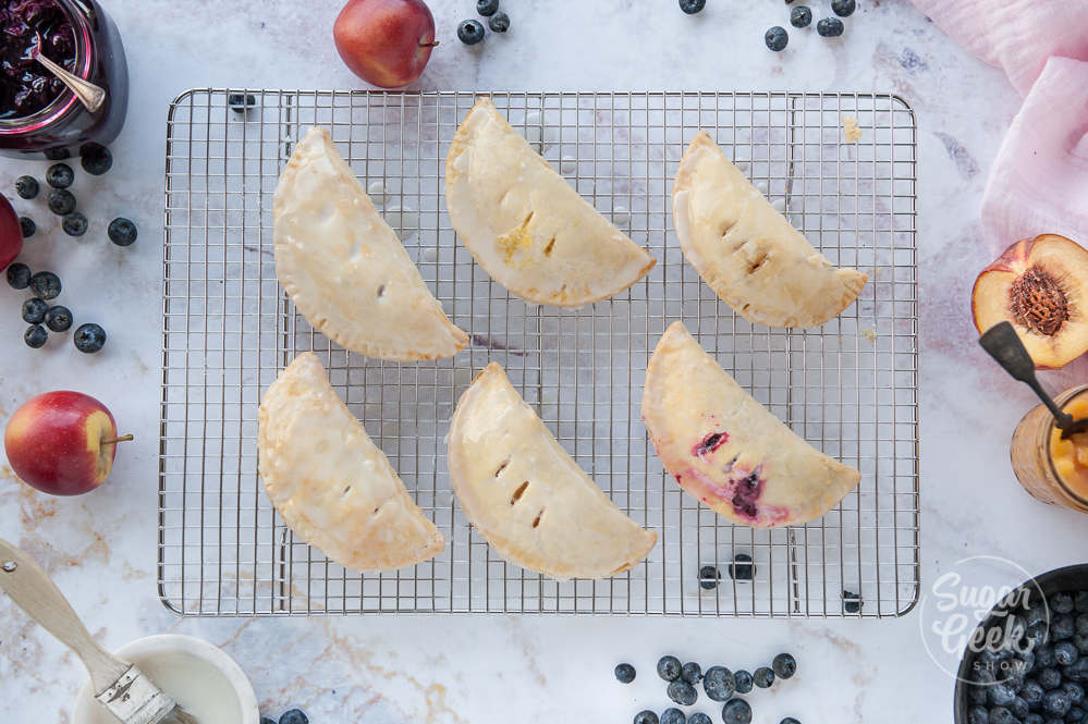 hand pies on a cooling rack shot overhead with a white backdrop and fresh fruit surrounding the edges