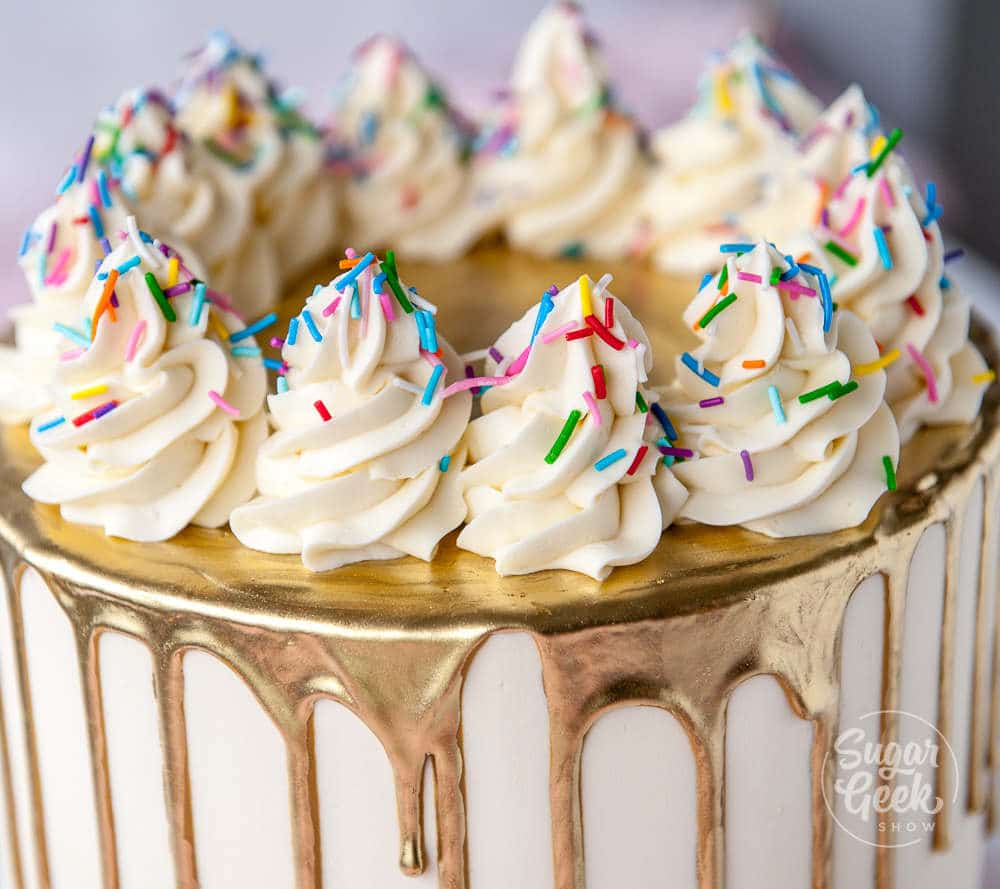 Close up of a cake with a gold drip, piped frosting on top and sprinkles.