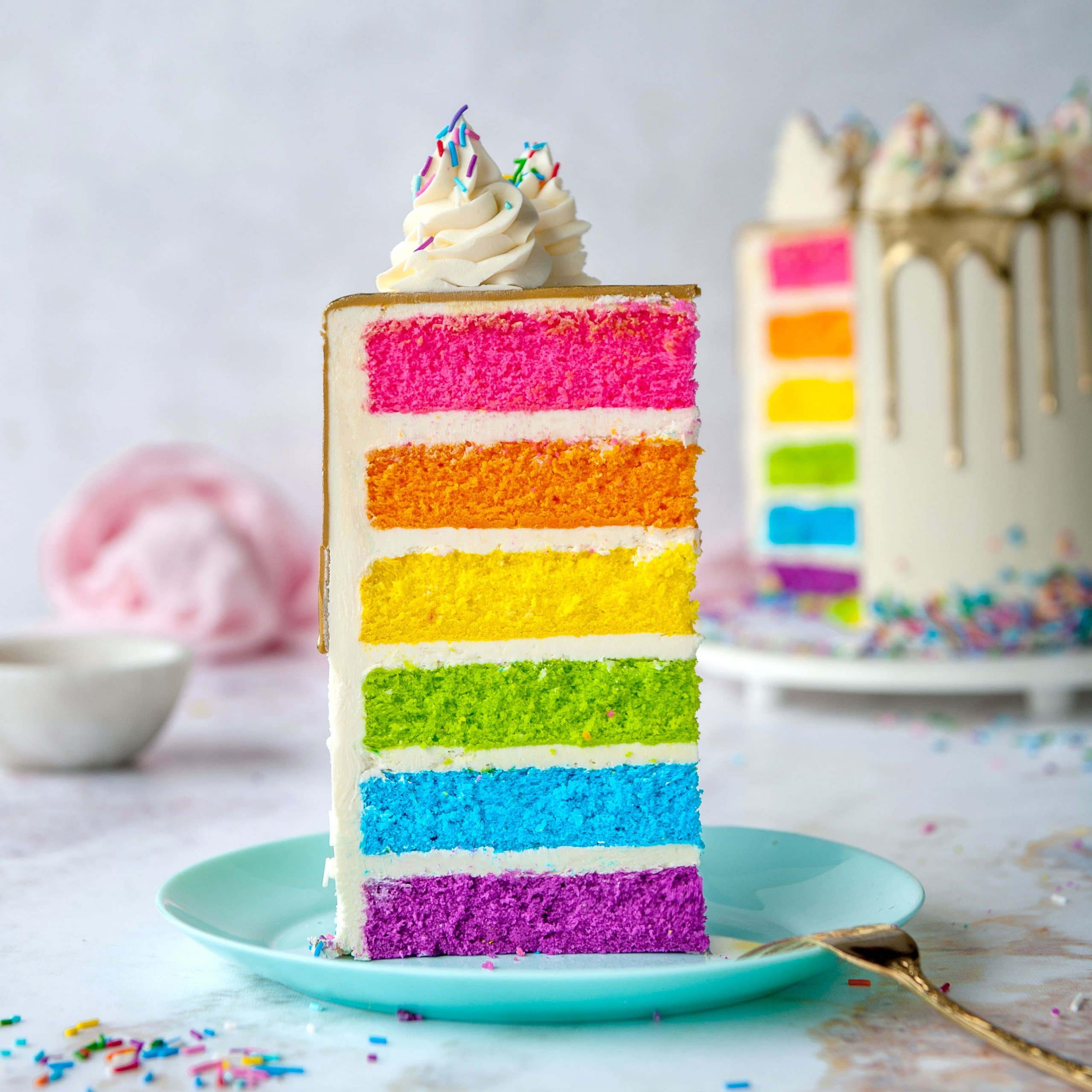 slice of rainbow cake on blue plate white background and whole cake in background
