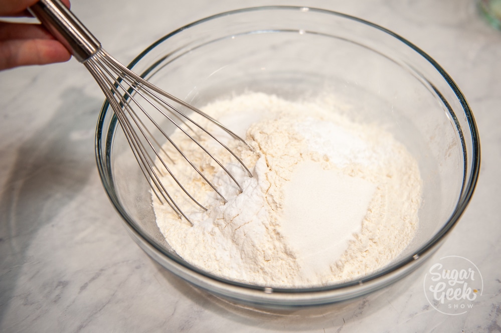 flour, sugar, salt, baking powder, baking soda in clear bowl with metal whisk