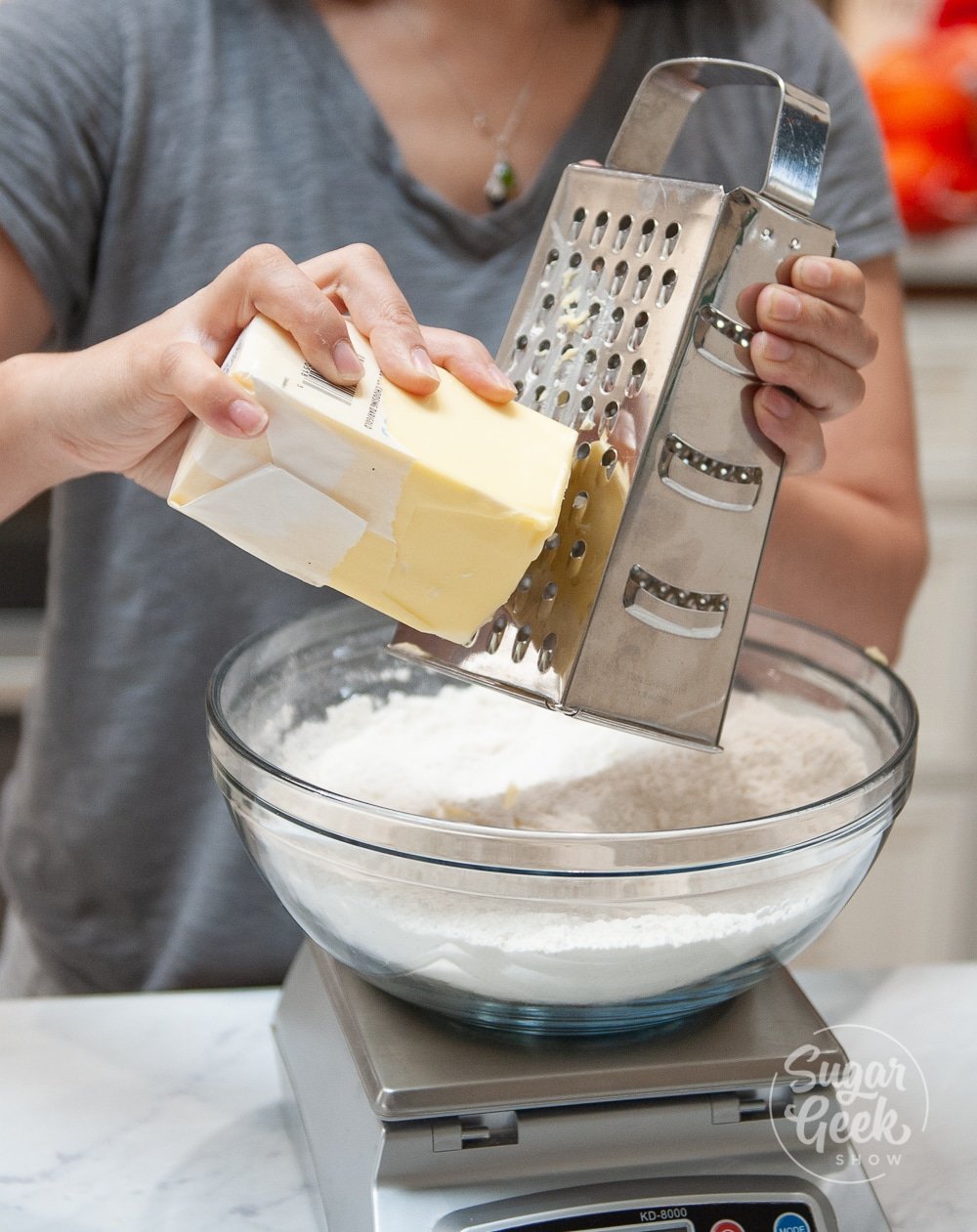 Woman grating cold butter into a bowl of dry ingredients sitting on top of a kitchen scale