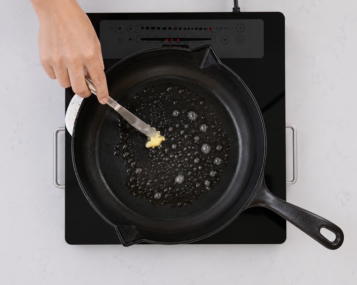 Hand melting butter with a butter knife in a hot cast iron pan on an electric stovetop.