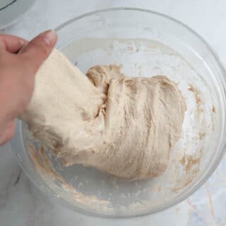 Hand pulling sourdough dough out of a clear glass bowl.