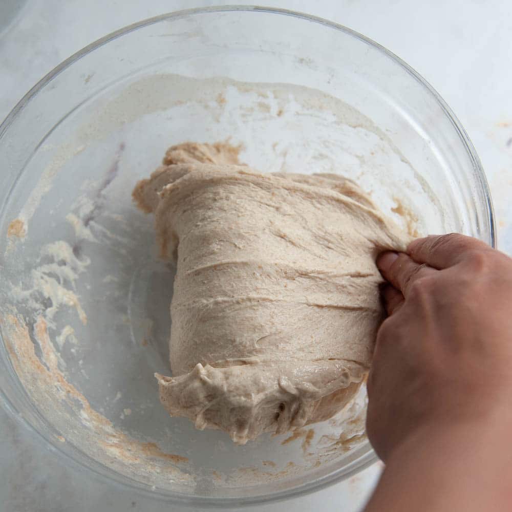 Hand folding sourdough dough over in a clear glass bowl.