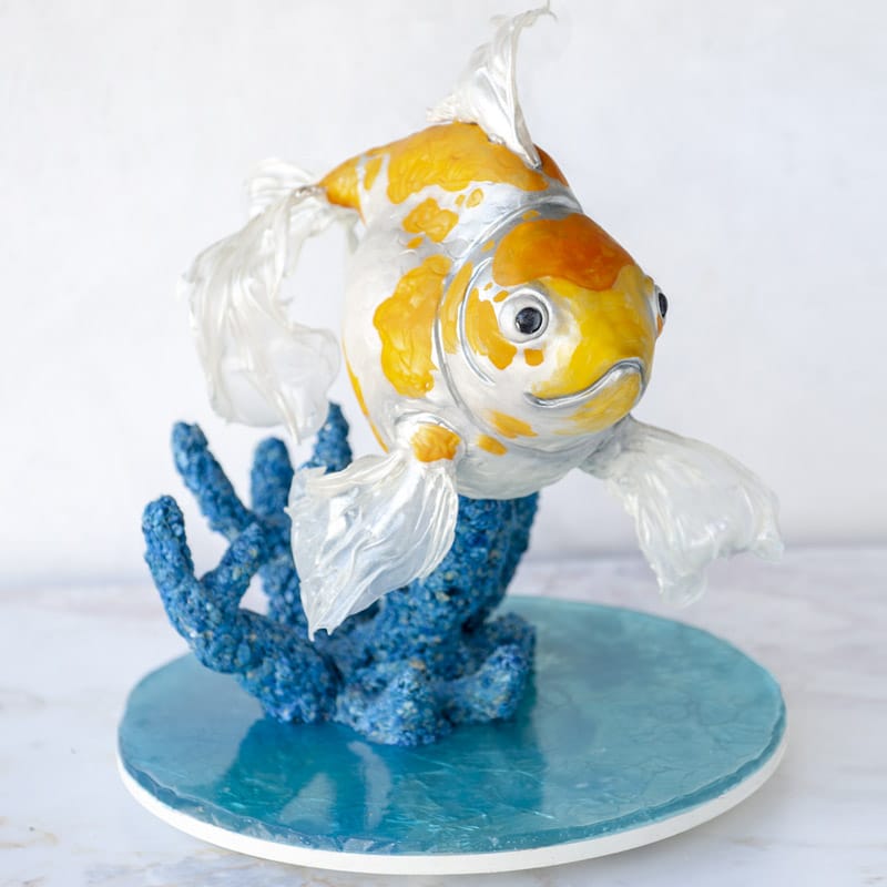 Cake sculpted into the shape of a goldfish with edible coral and a shiny isomalt cake board