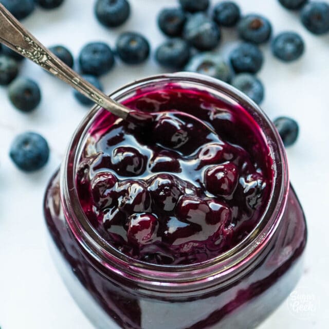 closeup of blueberry filling in a glass jar with a spoon and blueberries in the background
