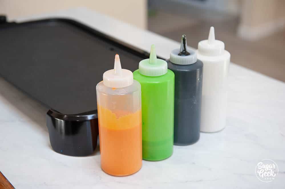 different colored pancake batter in squeeze bottles in front of a griddle
