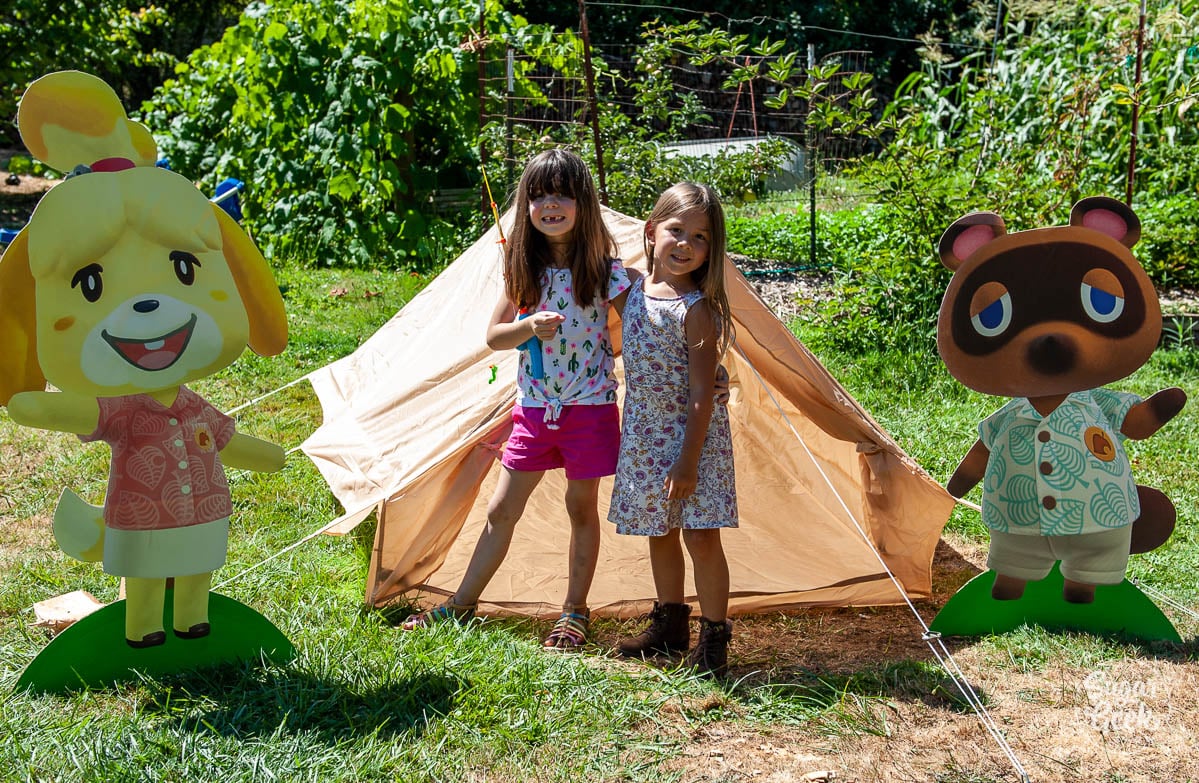 two girls standing in front of a tan a-frame tent with isabelle and tom nook on the sides