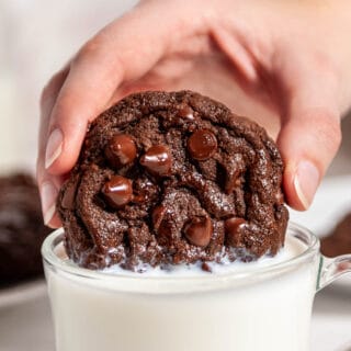 double chocolate chip cookie held by a hand close up being dunked in a glass mug of milk