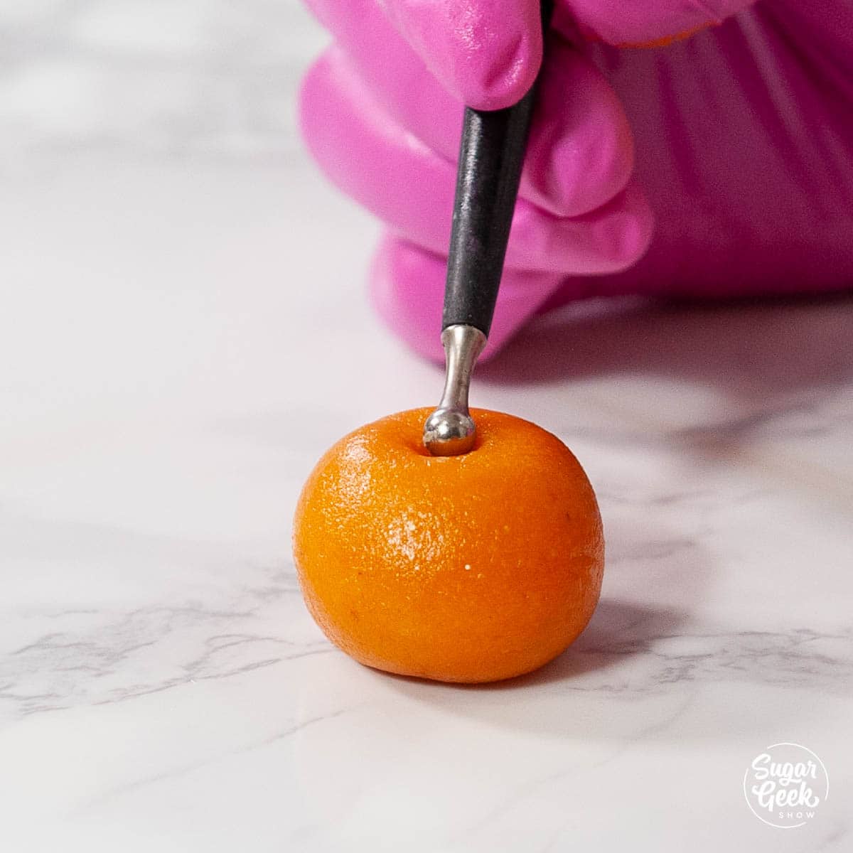 orange marzipan candy ball with pink gloved hand holding modeling tool