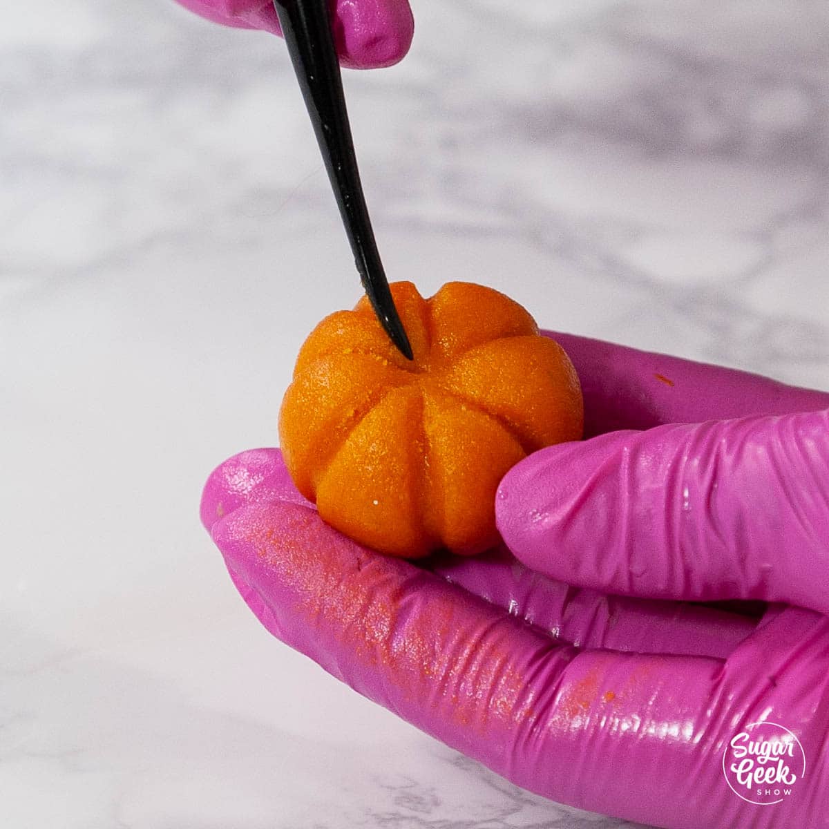 marzipan candy pumpkin held by pink gloved hand and black modeling tool above