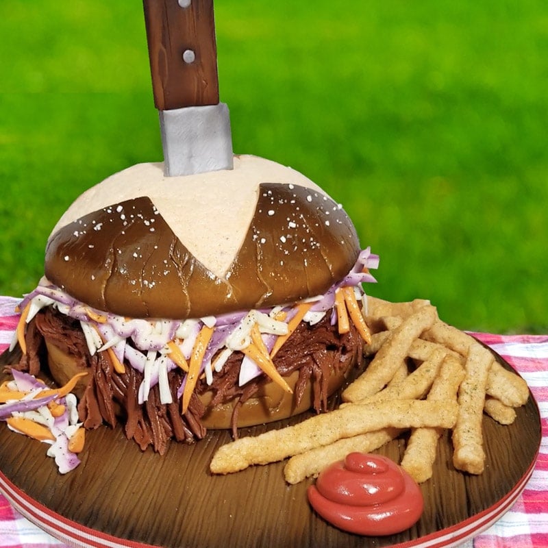 cake sculpted to look like a pulled pork sandwich with french fries, ketchup and a pretzel bun