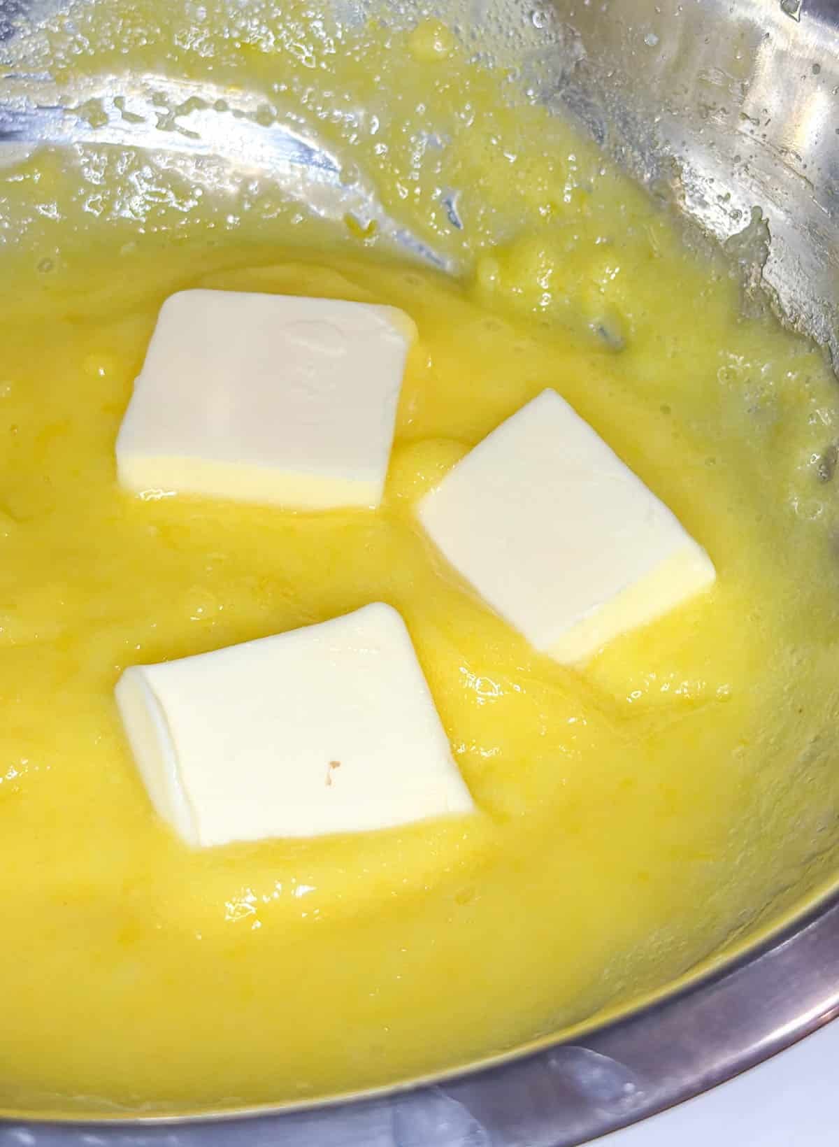 thickened lemon curd in a metal bowl with three cubes of butter