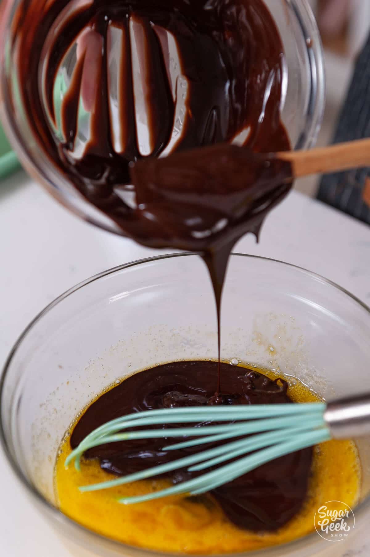 bowl of chocolate being poured into egg mixture with a blue whisk in front