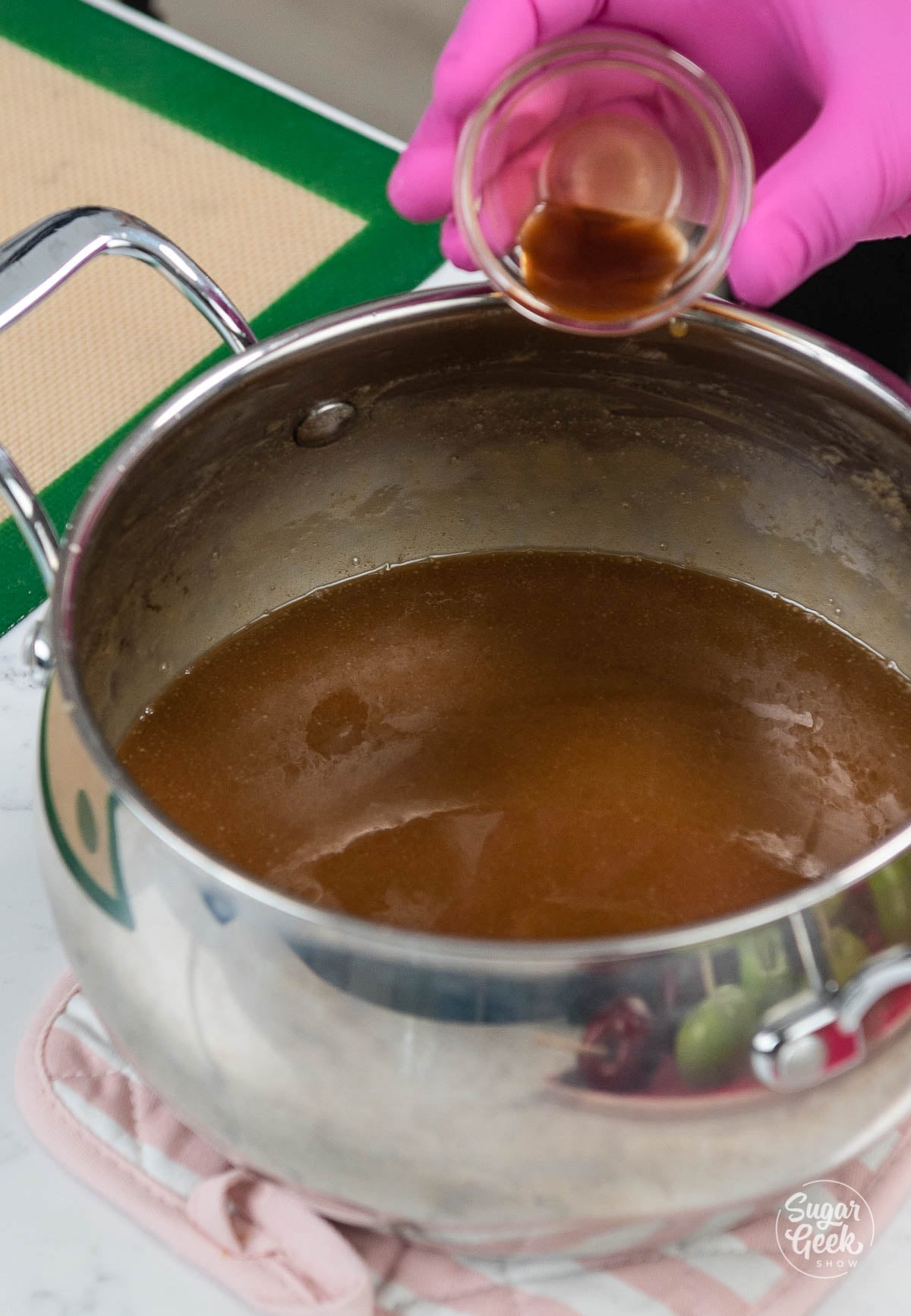 pink gloved hand pouring vanilla into a pot of cooked caramel
