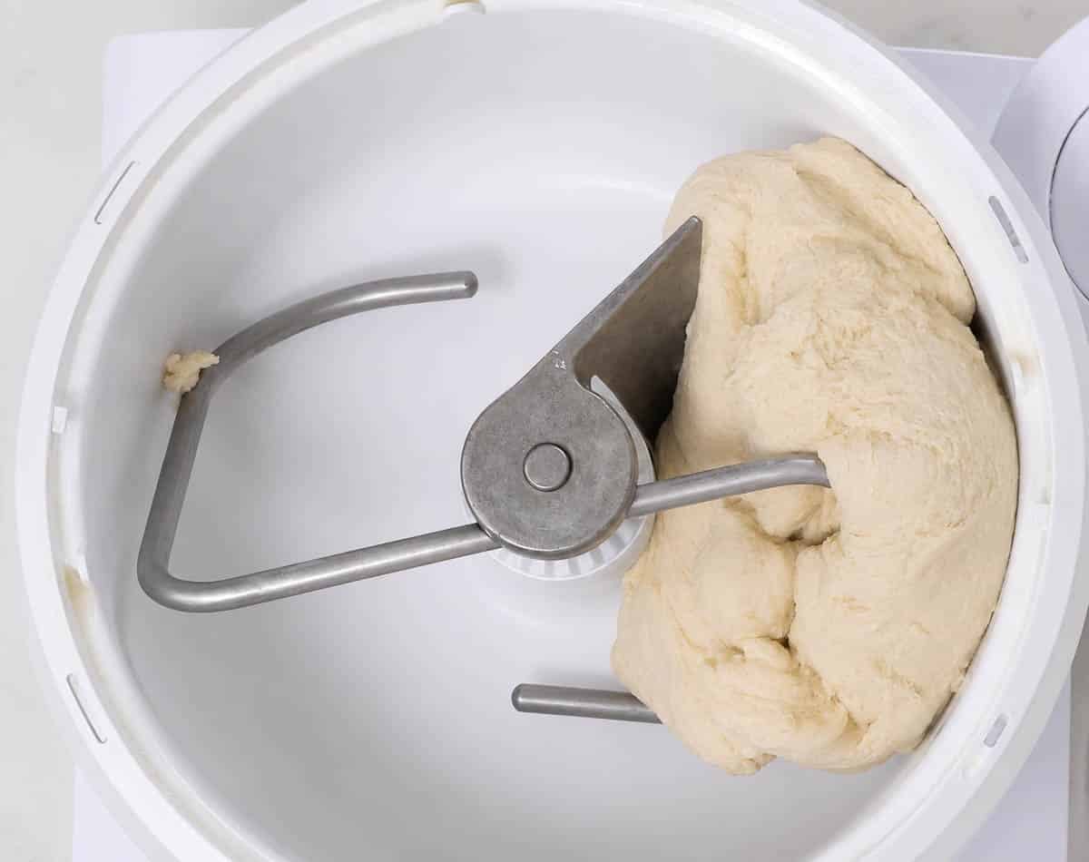 Bread dough in the bowl of a stand mixer with the dough attachment.