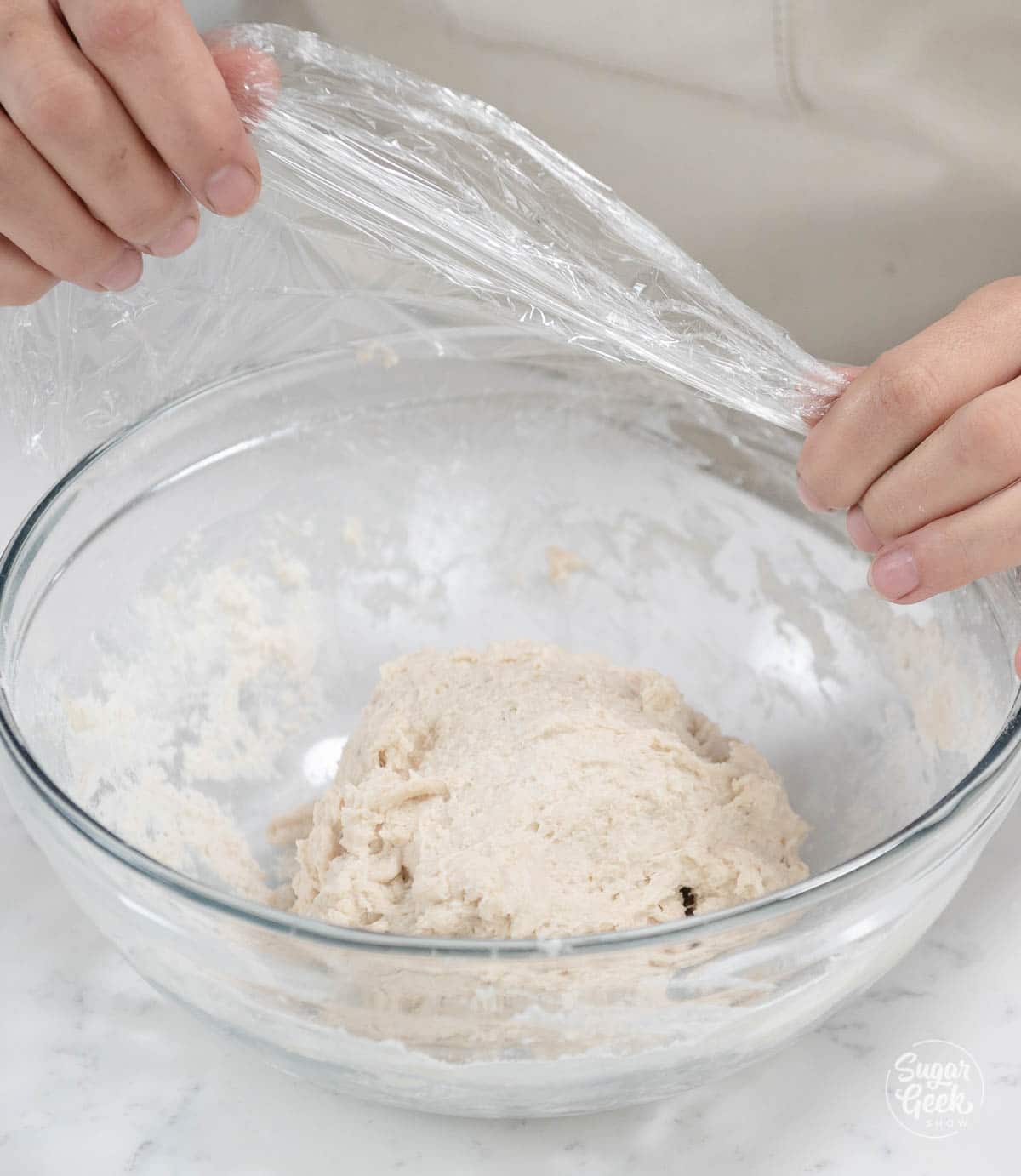 hands holding plastic wrap over dough in a clear bowl