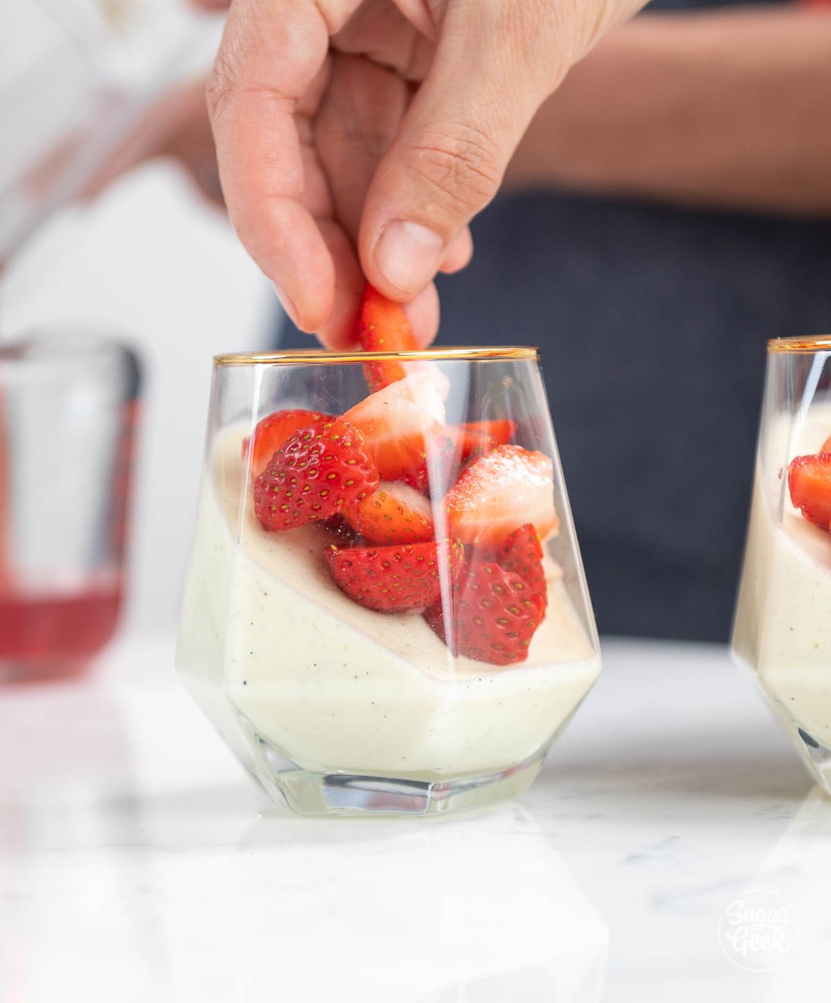 hand shown dropping strawberry pieces into wine glass filled with panna cotta